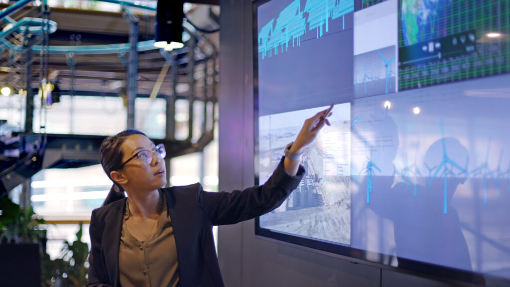 Stock photograph of a green card worker conducting a seminar / lecture with the aid of a large screen. The screen is displaying data & designs concerning low carbon electricity production with solar panels & wind turbines. These are juxtaposed with an image of conventional fossil fuel oil production.
