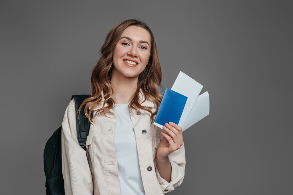 study abroad concept. student girl holding backpack, book, notebook, passport isolated on a dark grey background, copy space, immigration, student visas